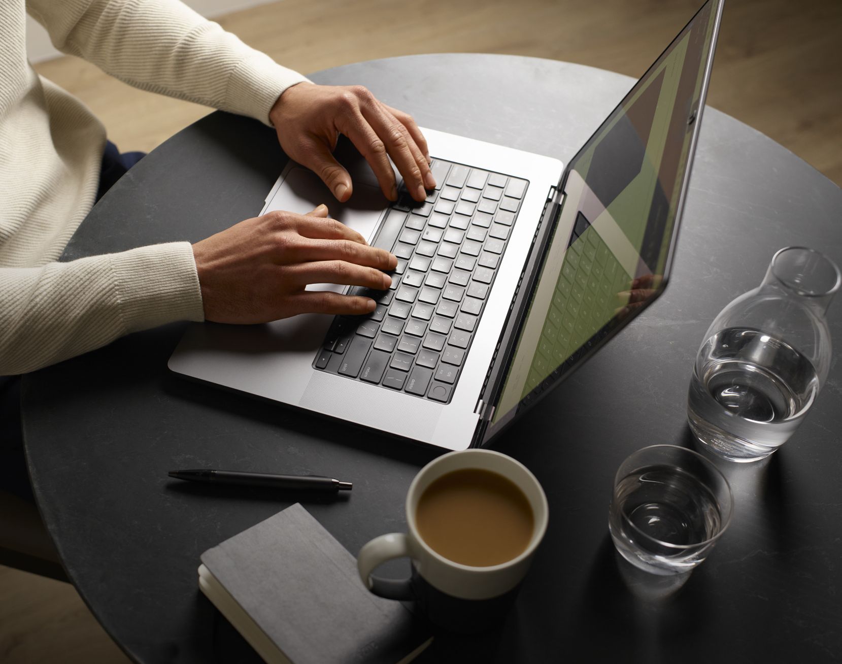 Hands typing on a laptop on a coffee table. There is a pen and warm cup of coffee on the table.