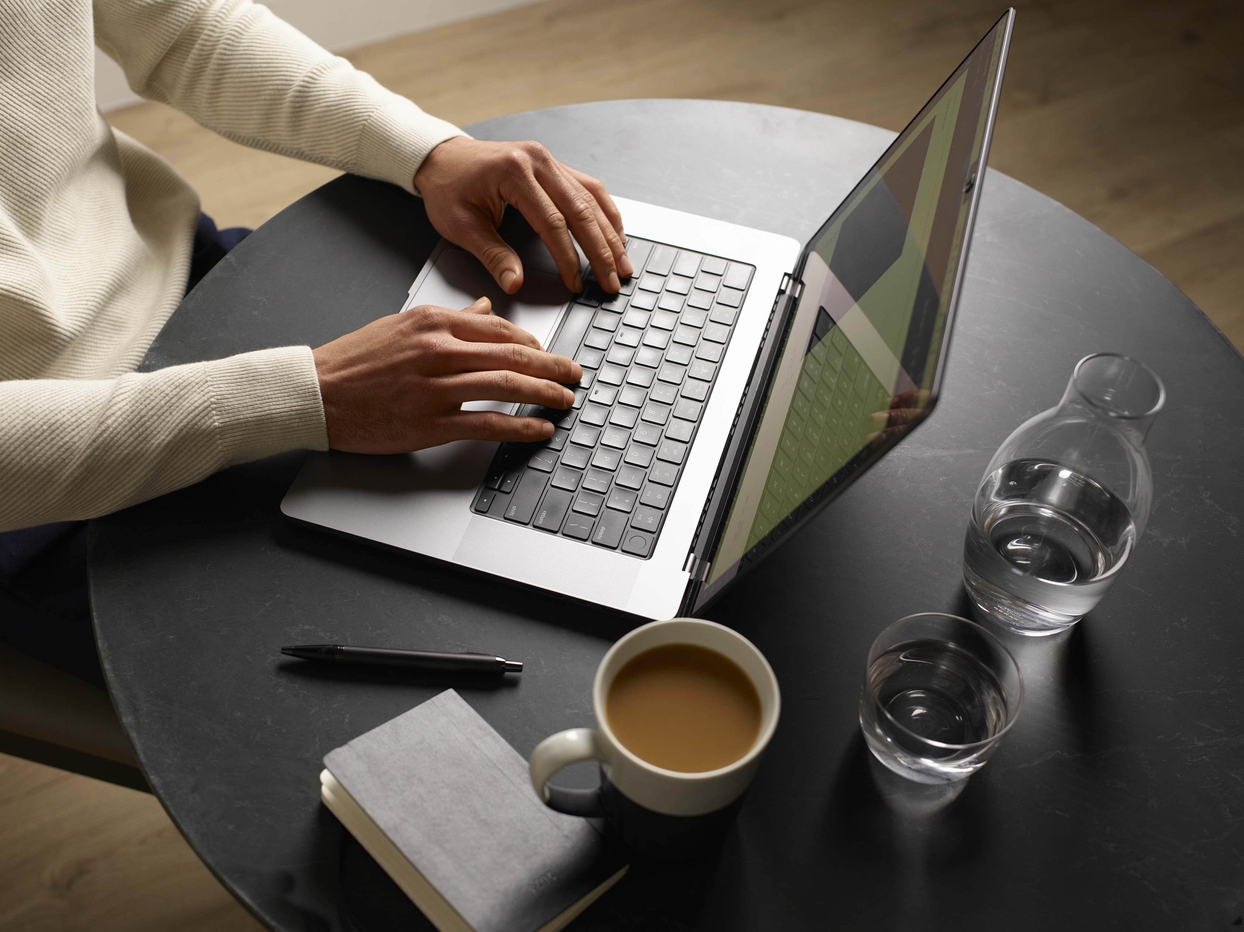 Hands typing on a laptop on a coffee table. There is a pen and warm cup of coffee on the table.